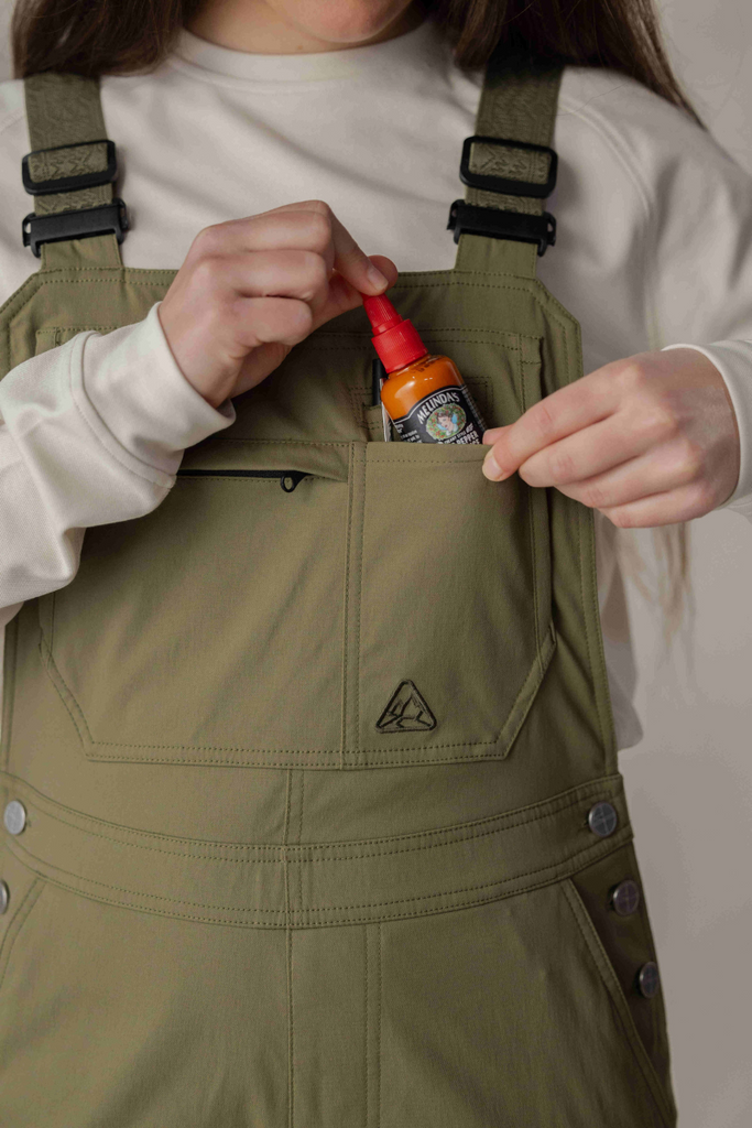 Model putting bottle of hot sauce in front pocket of Ecotrek overalls in Olive